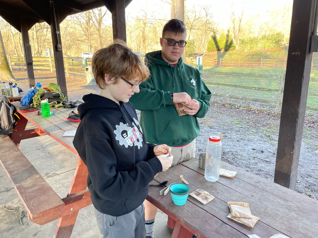 Two scouts prepare their oatmeal breakfast by pouring mix into a cup.