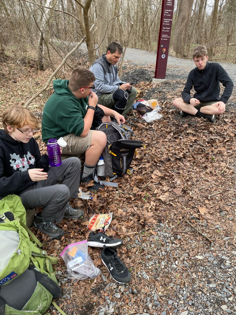 Several scouts sit on a log alongside a trail for lunch.