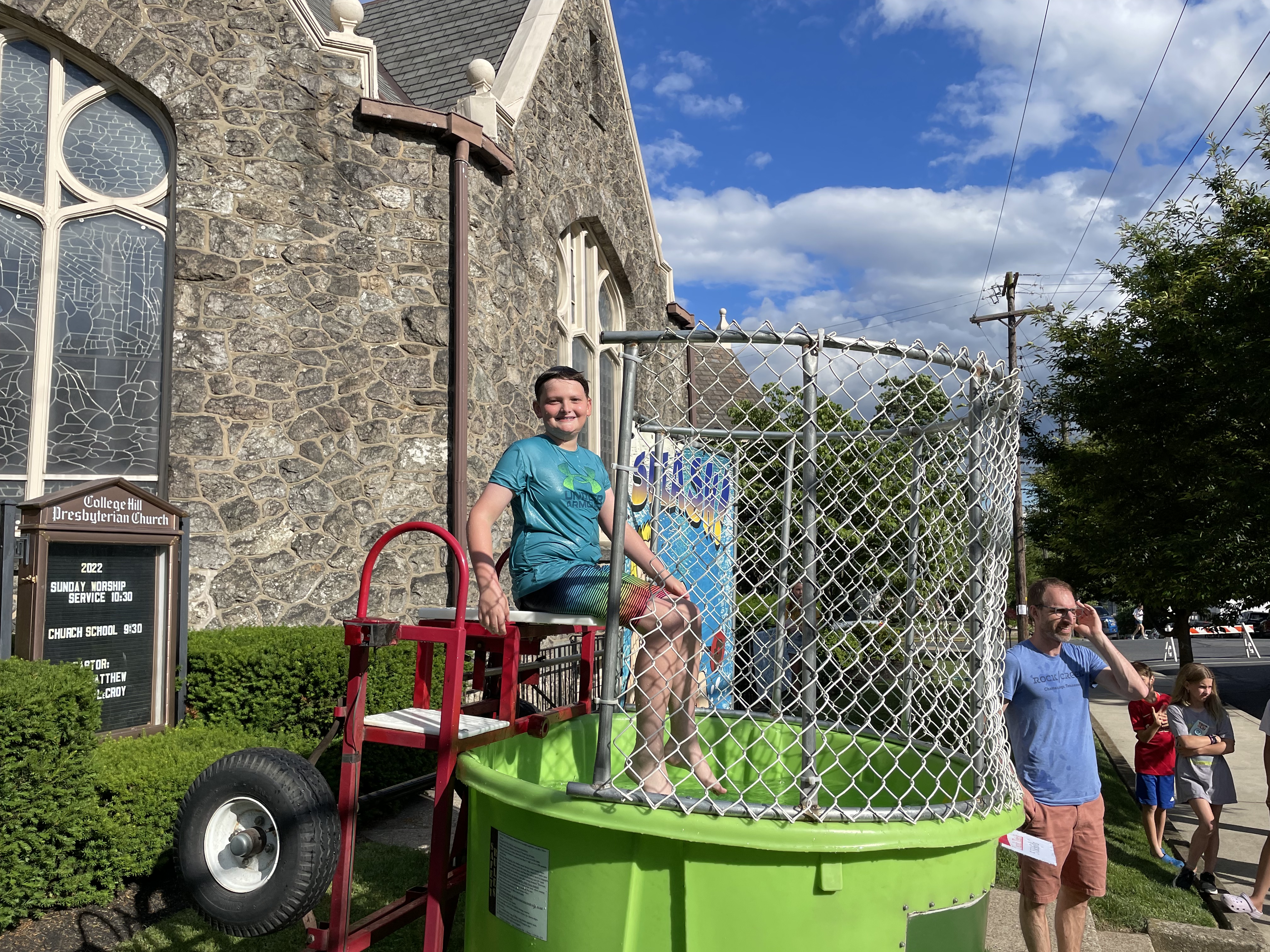 A scout in a blue shirt sits in a dunk tank. A church can be seen behind him.